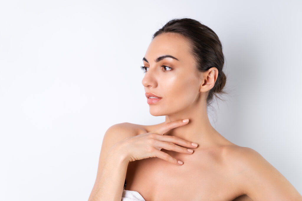 A portrait of a young woman with glowing skin, her hair tied back, touching her neck and shoulder. She is looking to the side with a calm expression after getting a Photofractional facial.